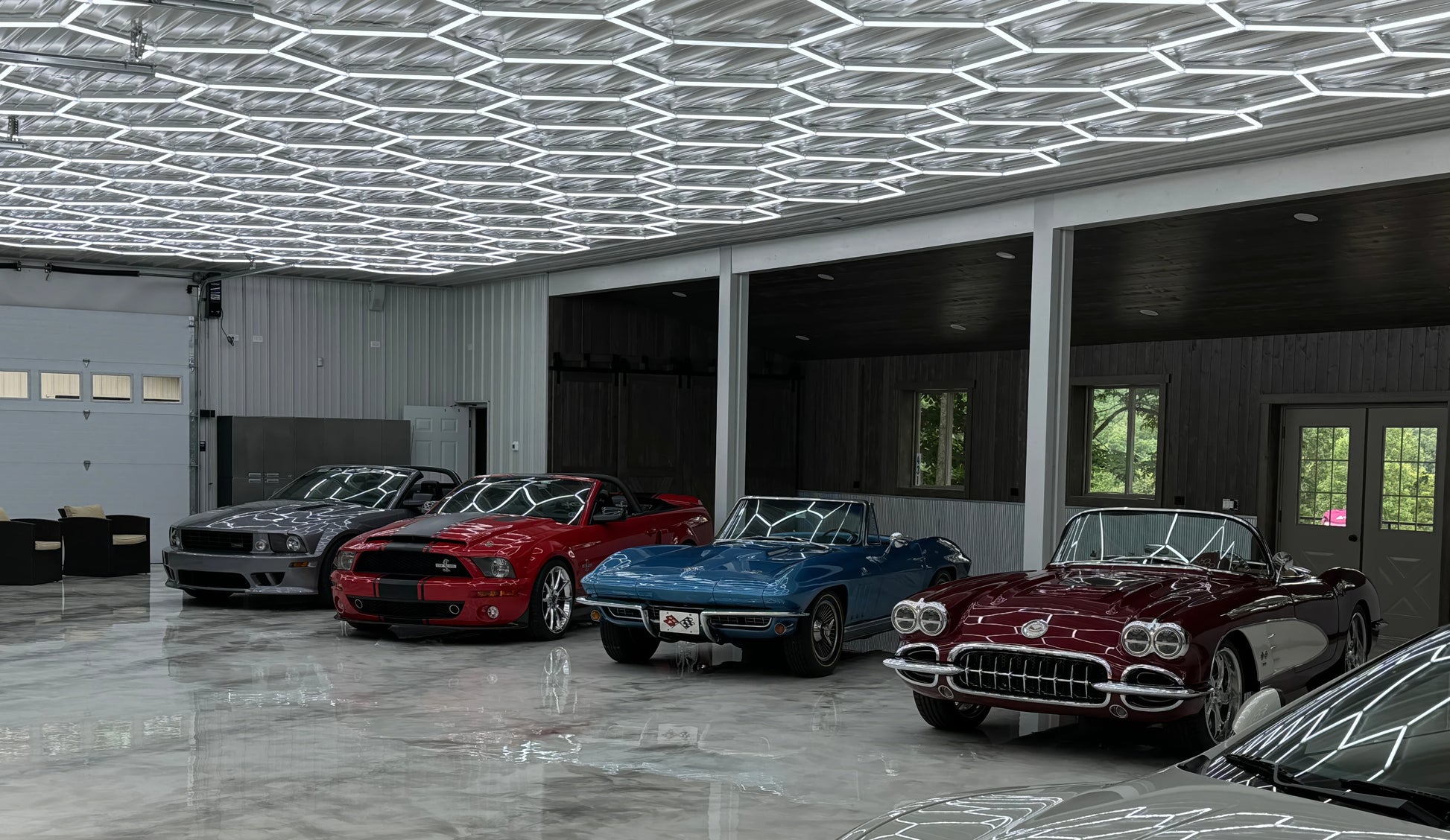 A row of classic cars parked inside a spacious modern garage with a geometric honeycomb LED ceiling light and polished concrete floors. The cars include a silver classic car, a red muscle car, a blue convertible, and a dark red convertible with chrome details.