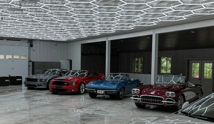 A row of classic cars parked inside a spacious modern garage with a geometric honeycomb LED ceiling light and polished concrete floors. The cars include a silver classic car, a red muscle car, a blue convertible, and a dark red convertible with chrome details.
