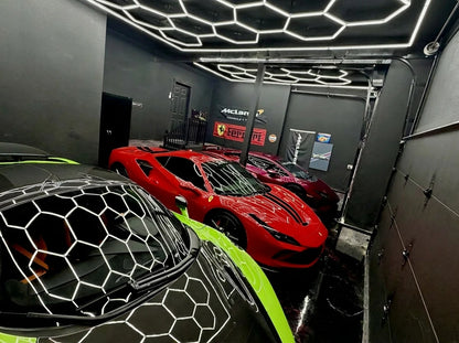 A high-end garage or showroom with dark gray walls and a distinctive hexagonal LED light grid on the ceiling. In the foreground, the hood of a black sports car with neon green accents is visible, reflecting hexagon ceiling lights.