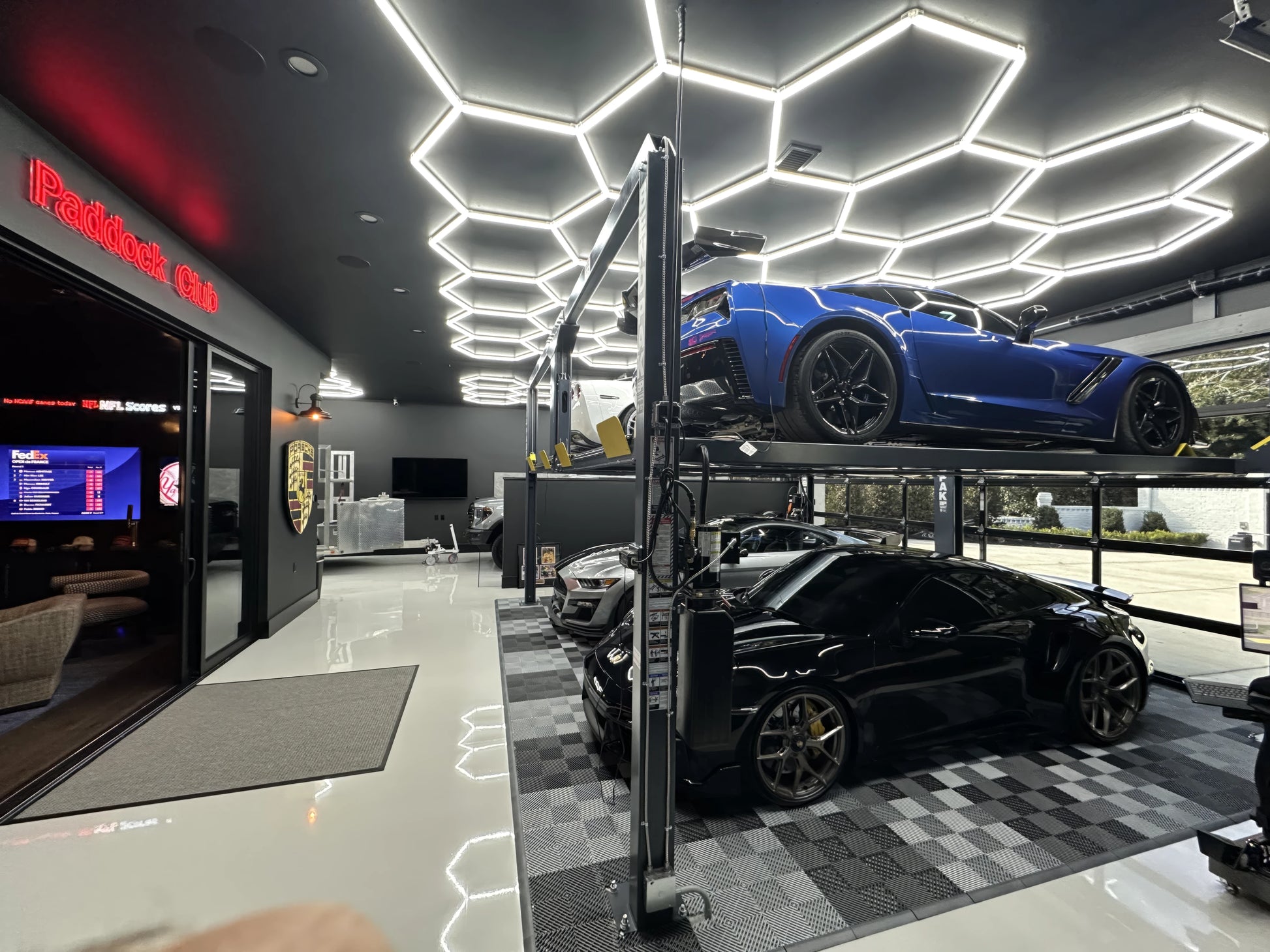 A luxury home garage featuring a blue sports car elevated on a two-post car lift and a black sports car underneath on a checkered floor mat. The garage is equipped with a modern ceiling of white hexagonal LED lights, a dark gray wall with a red 'Porsche Club' sign, and a Porsche crest on the wall.