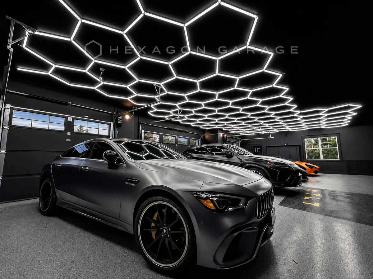 A matte dark gray Mercedes-AMG sports car parked inside a modern garage with a large hexagonal LED ceiling light installation. The brand name 'HEXAGON GARAGE' is watermarked across the top center.